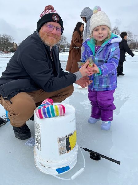 A young girl standing on a frozen lake smiles for a photo while her father, kneeling by her side, holds a bluegill just caught through the ice.
