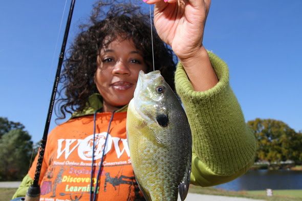 An adult woman in the background holds a fish on the end of a line up toward the camera.