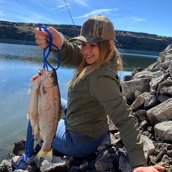 A girl sitting on a lake banks girl smiles while holding two large trout.