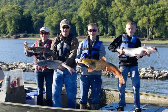 A family of a mother and father and two boys each holds a large catfish caught on a trotline as they stand in a boat on the river.