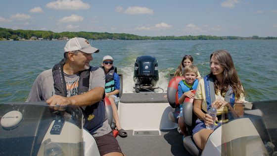 A family of five, two adults and three children, wearing life jackets and smiling while boating on a sunny day.