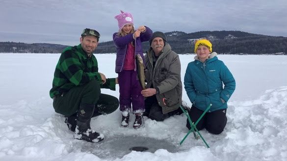 A family of four fishing on ice with a tip-up in the foreground, a child is holding the lake trout