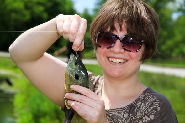 A close-up shows a woman holding up a fish she just caught and smiling.