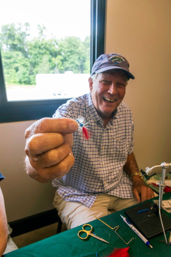 A fly tier holds up a fly toward the camera and smiles, with fly tying equipment seen on a table in front of him.