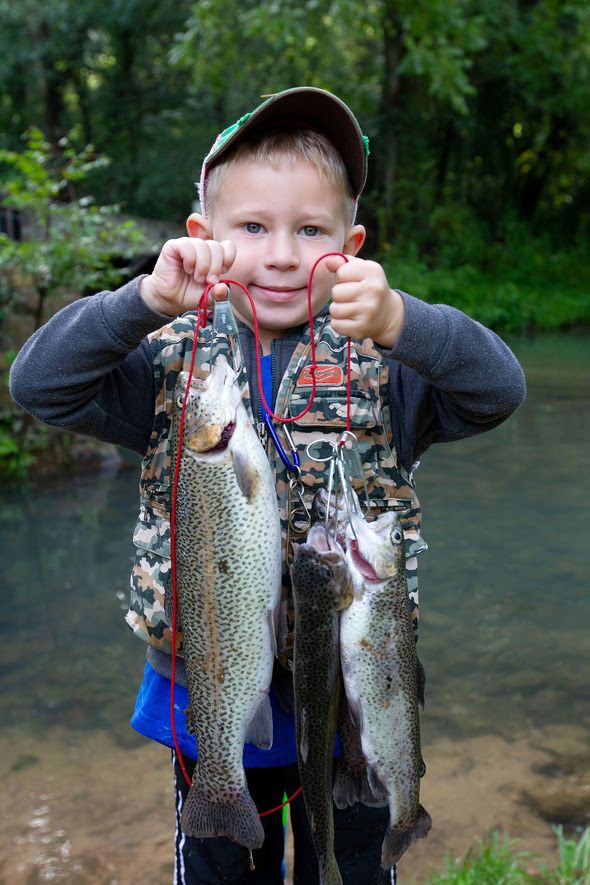 A young boy holds up three trout on stringers that he just caught while beaming proudly.