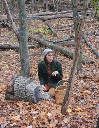 woman in a hat and jacket, holding mushrooms, sits on ground next to tree stumps in a forested area