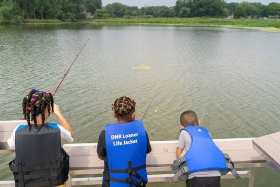 Three children, all wearing life jackets, fishing off a pier on a pond.